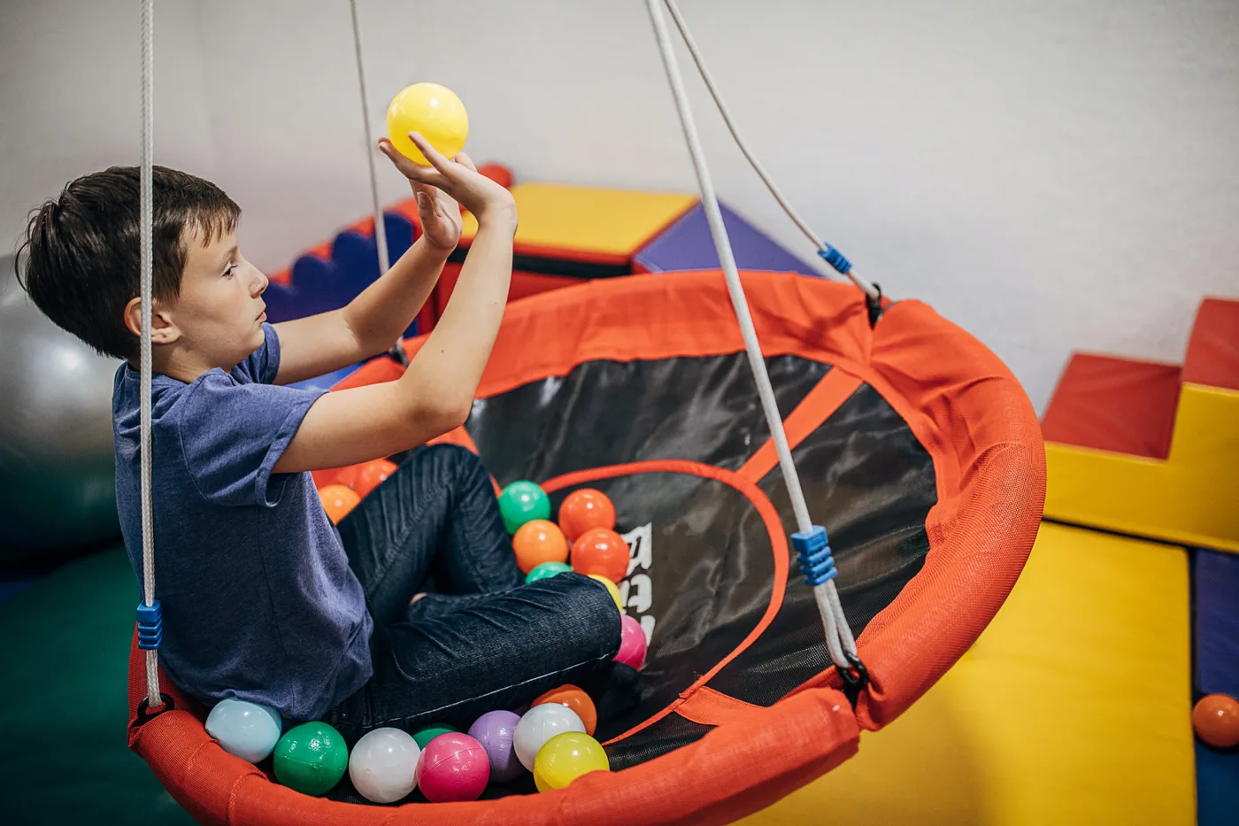 Menino em balanço de rede segurando uma bola, em sala de integração sensorial